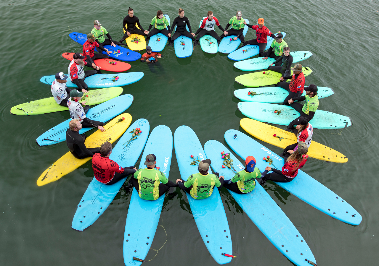 Operation Surf participants and veteran supporters sit on colorful surfboards in a circle on the water, holding hands during a healing paddle out surf session