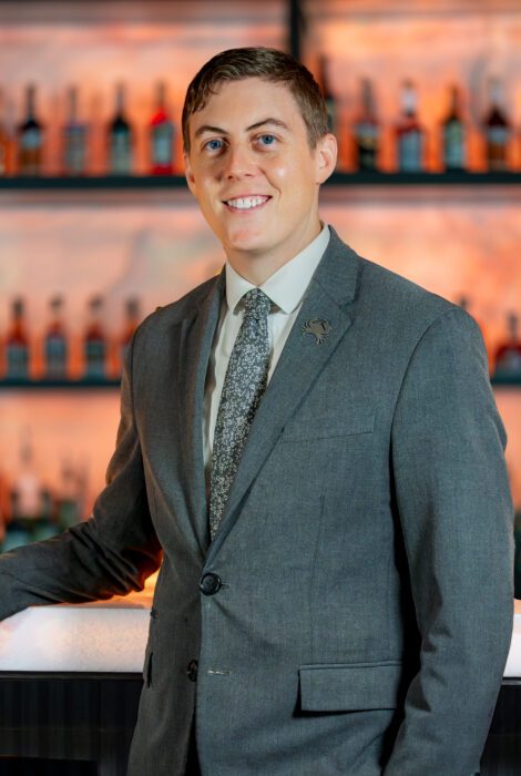 service manager Alex Rediske in a gray suit with shirt and tie smiling in front of the bar with the back bar lit and filled with liquor bottles
