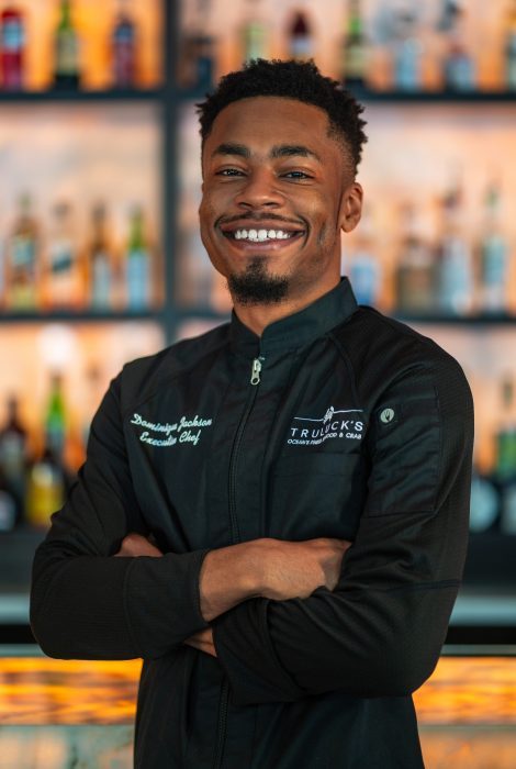 Truluck's Arboretum executive chef Dominique Jackson in a black chef coat with his arms folded across his chest in front of the back bar