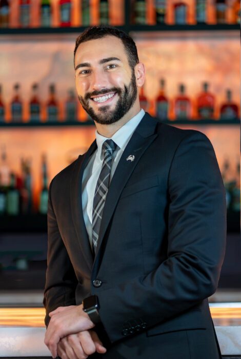 headshot of Cameron Stefanini, beverage manager of Truluck's Austin with his elbow leaned onto the bar in a dark navy suit with shirt and tie, smiling in front of the bar top
