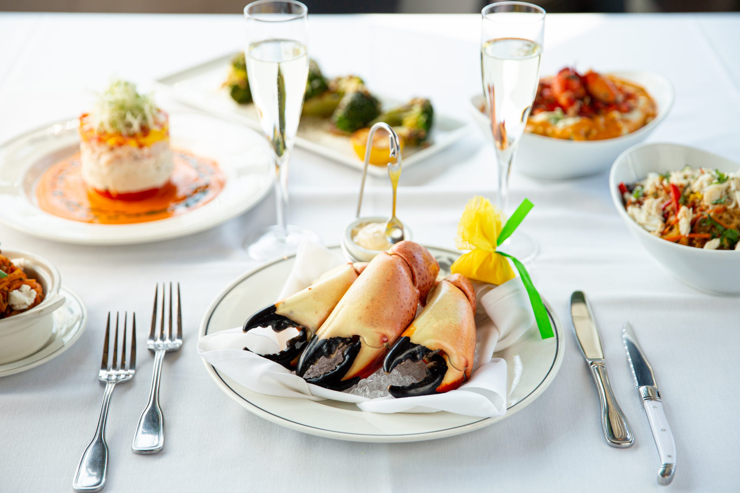 picture of Florida stone crab claws on a plate with a lemon wrap and sides of broccoli, lobster mashed potatoes and crab and shrimp Napoleon in the background