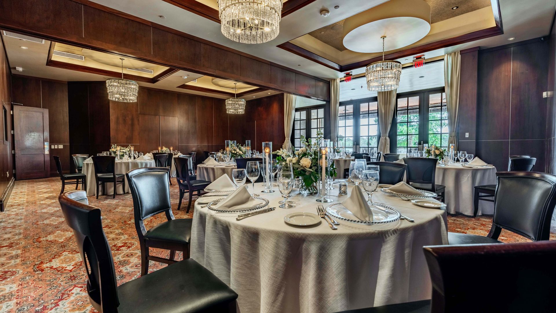 The Stone Crab Room at the Woodlands location. Corner viewpoint of round tables dressed in white linen, china, glass and silver with large glass French doors overlooking the terrace.