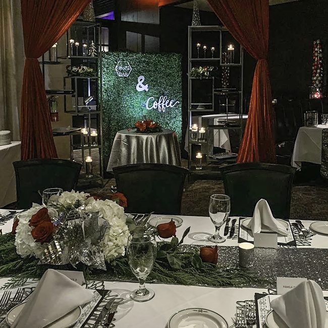 picture of Truluck's Woodlands private holiday event with red, white and green accents. Tables are covered with a silver runner and floral centerpieces to include holiday greenery, red roses and white hydrangea. In the background is a dessert and coffee station setup with tiered shelving for guests to choose a dessert and be served a beverage.