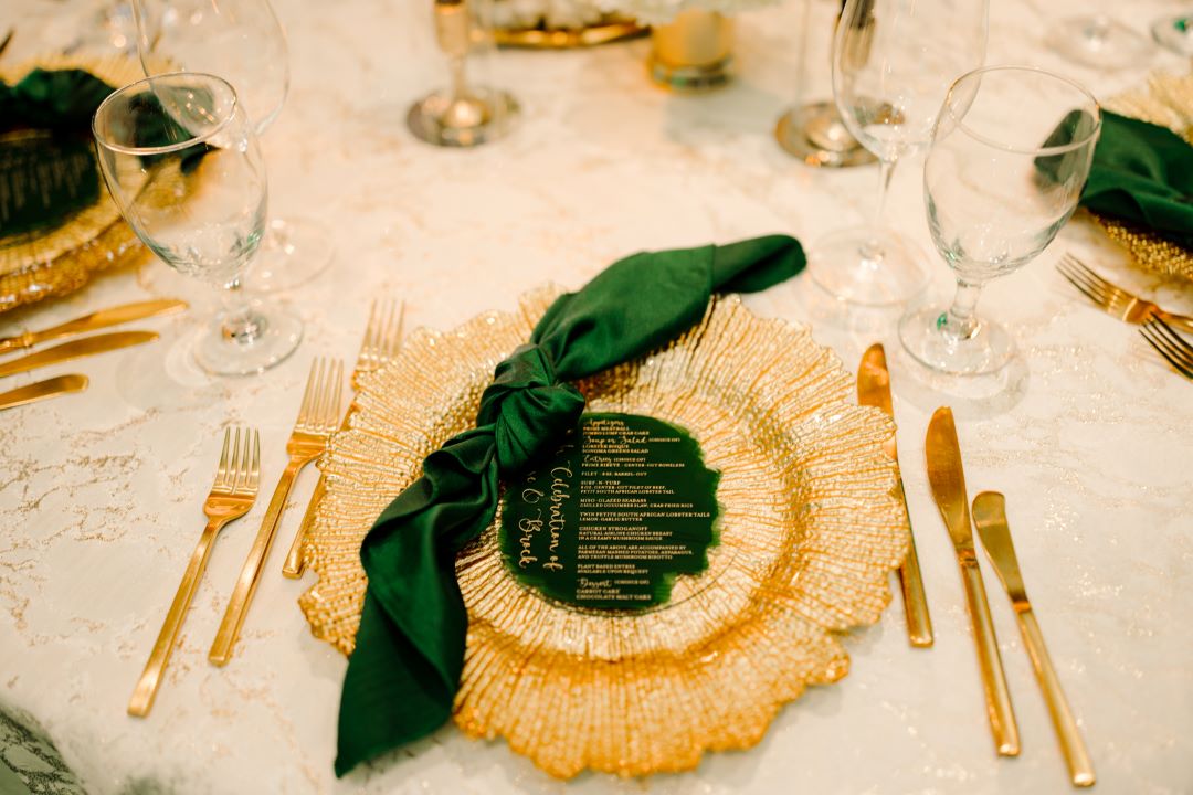 a gold charger atop a white and gold tablecloth accented with gold silverware and a custom emerald green menu with emerald green napkin tied in a knot