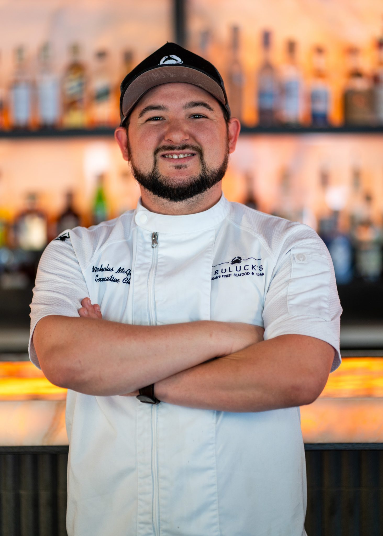 Truluck's Austin downtown executive chef Nick McGuire in a white chef coat with his arms folded on this chest in front of a backlit bar top