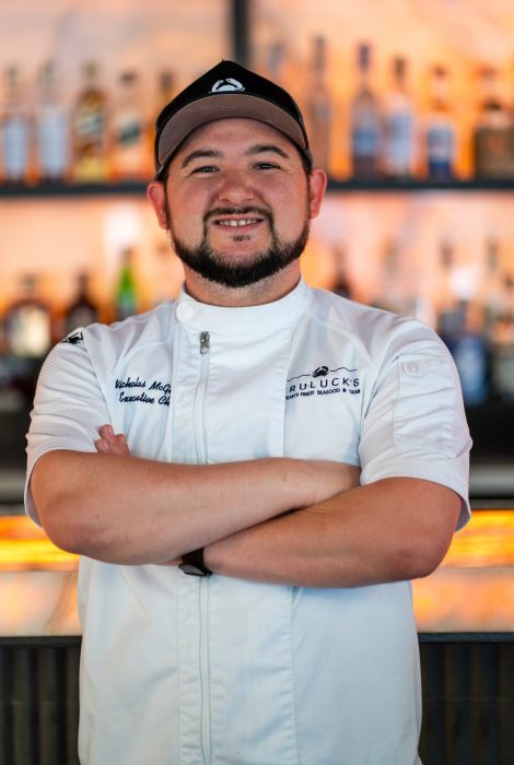 Truluck's Austin downtown executive chef Nick McGuire in a white chef coat with his arms folded on this chest in front of a backlit bar top