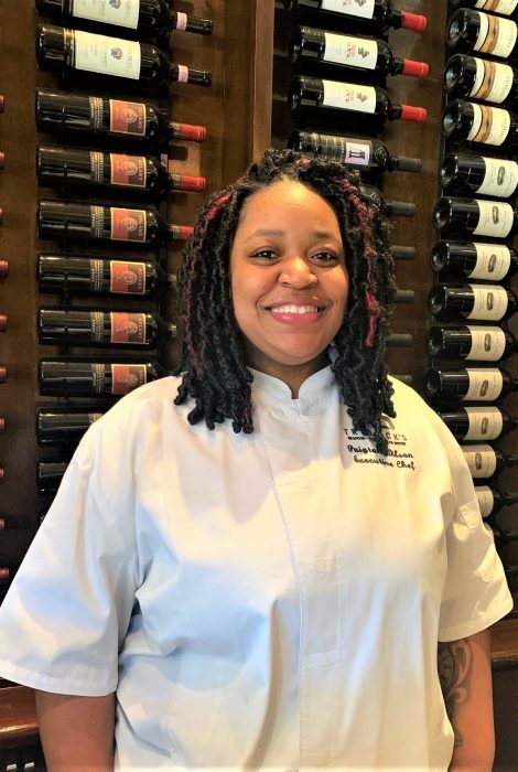 picture of Chef Paigton in front of the wine wall at the restaurant