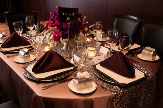 picture of Truluck's Woodlands private event with lavender table linen, satin eggplant napkins, silver table runner, silver chargers and take away boxes of chocolates. Florals are magenta and purple flowers.