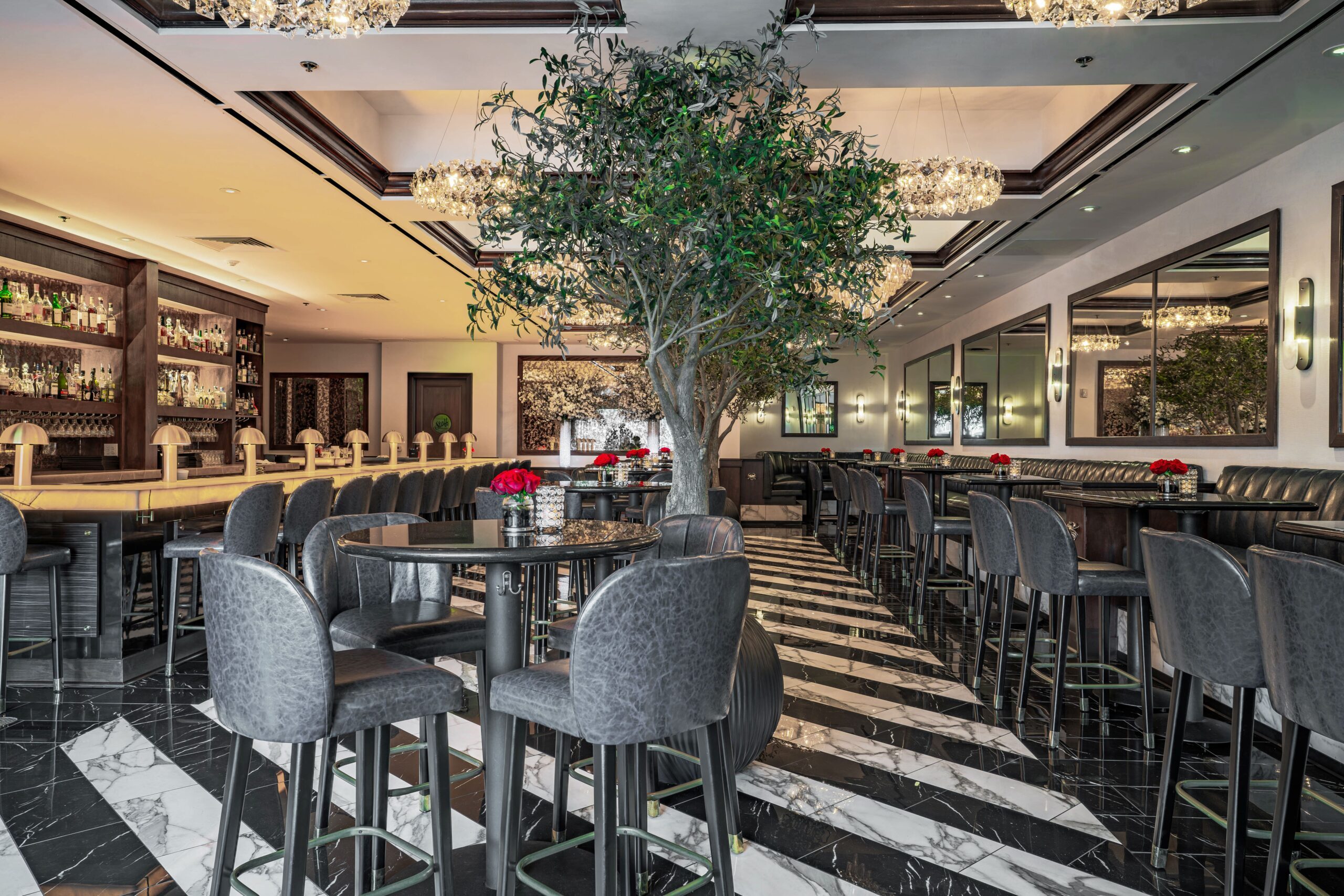Elegant bar and lounge at Truluck’s Houston featuring black marble flooring, gold chandeliers, indoor olive trees, and a full bar lined with leather seating.