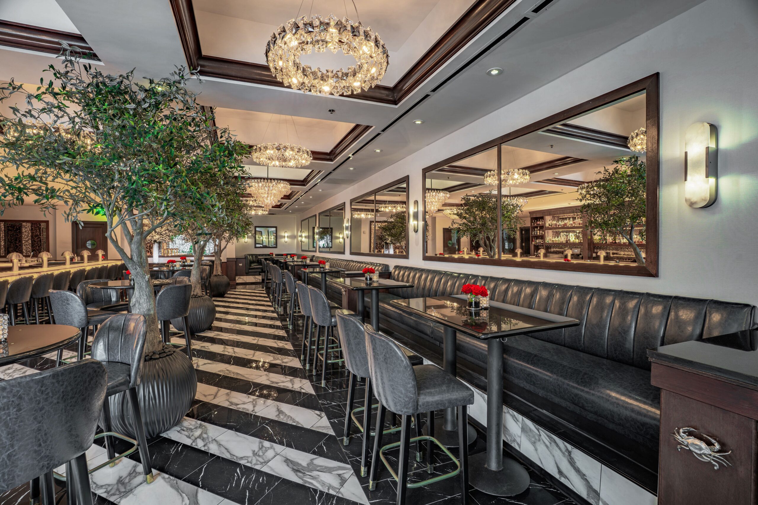 Upscale bar and lounge seating at Truluck’s Houston featuring black banquettes, marble flooring, crystal chandeliers, and indoor olive trees creating a refined, inviting atmosphere.