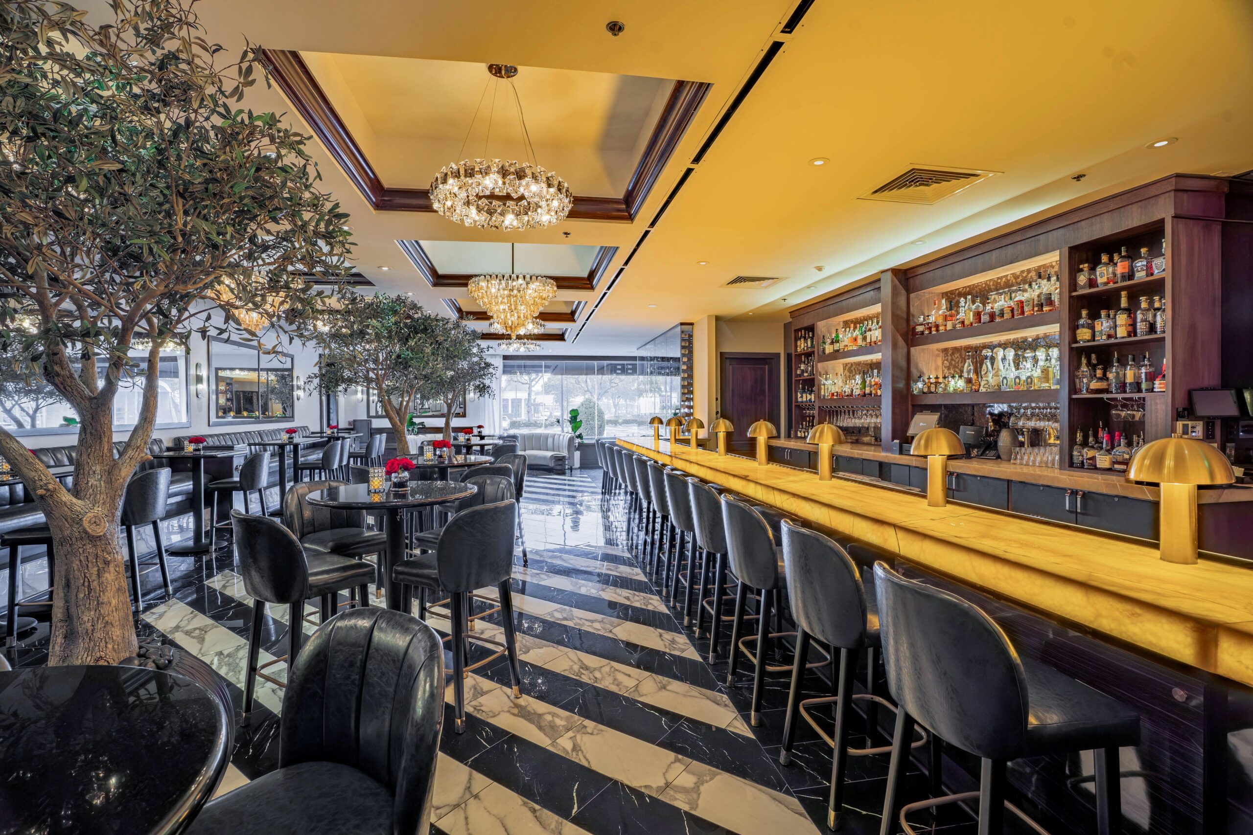 Luxurious bar at Truluck’s Houston with illuminated gold bar top, leather barstools, crystal chandeliers, and a premium backlit spirits display.