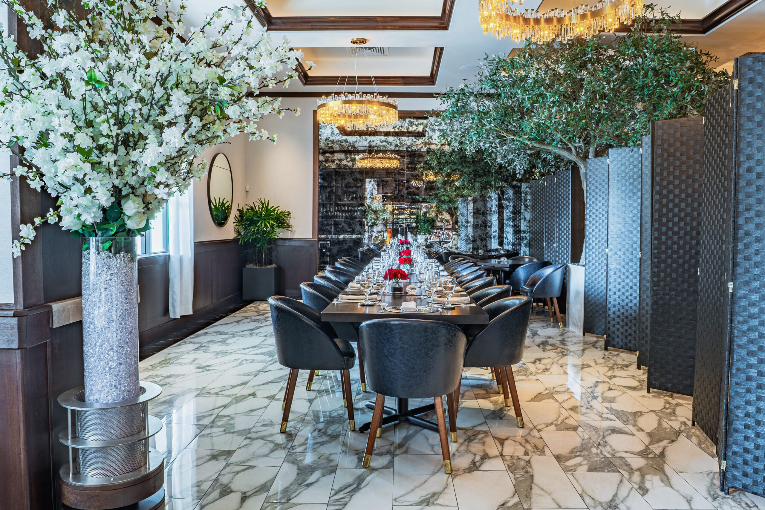Elegant group dining room at Truluck’s Houston called the TRU 1 room, featuring a long banquet table set with fine glassware and modern place settings, marble floors, lush indoor trees, and crystal chandeliers—ideal for upscale events, celebrations, and corporate dining.