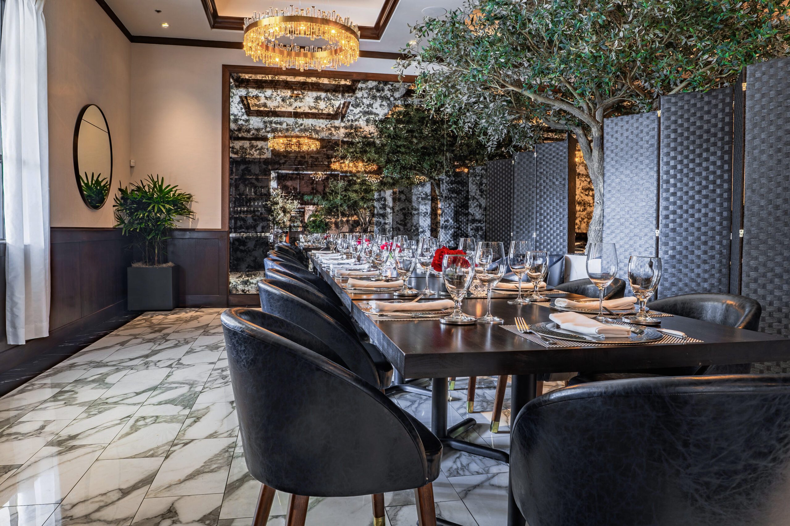 Upscale group dining room at Truluck’s Houston called the TRU 1 room featuring a long, elegantly set table with glassware and linens, marble floors, modern navy chairs, mirrored walls, and lush indoor trees under crystal chandeliers—ideal for luxury events and group dining.