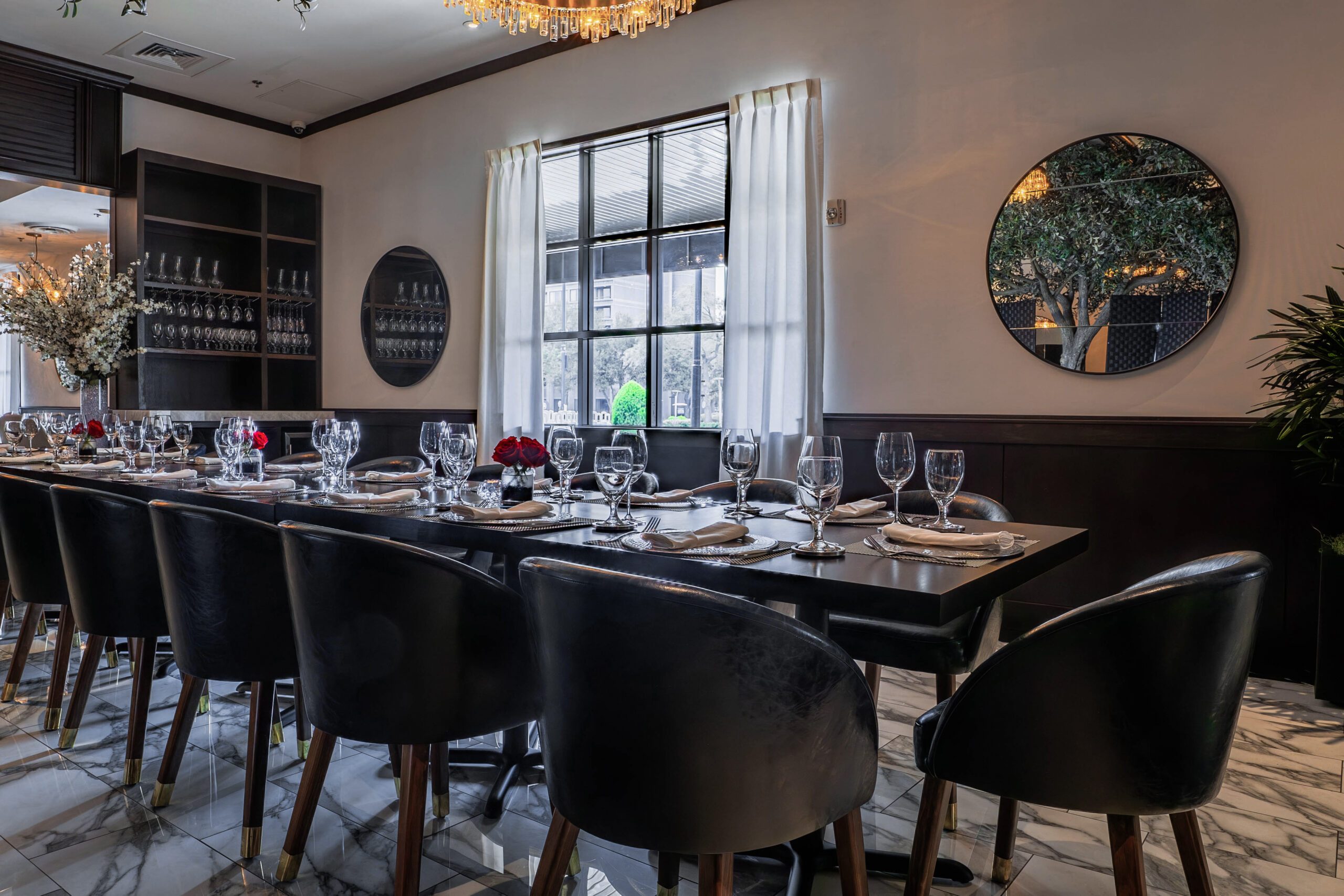 Elegant group dining space at Truluck’s featuring a fully set private event table, polished marble floors, chic modern seating, lush greenery, and warm chandelier lighting.