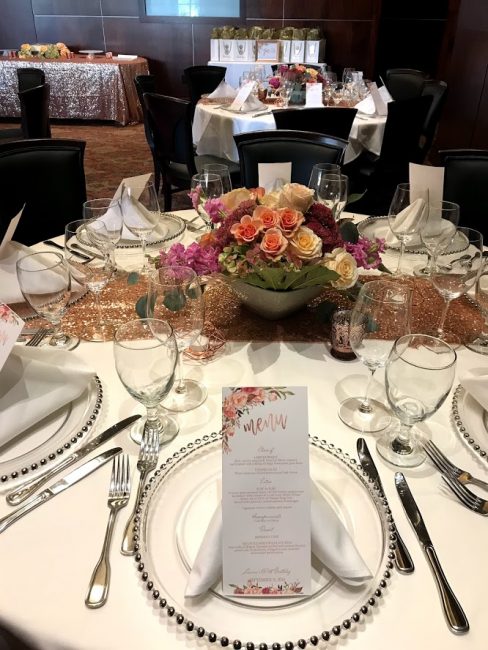 picture of Truluck's Woodlands private dining event for a shower with white linen, gold table runner and spring colored bouquet of flowers in the center. In the distance is a dessert table with a rose gold sequin linen and gift bags.