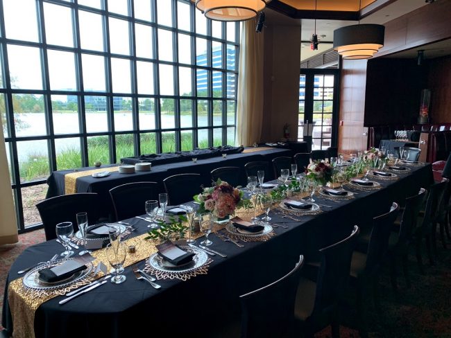 picture of Truluck's Woodlands private dining event with all black linen and gold accents. Table centerpieces are deep purple Dahlias with dusty rose colored roses. This is a long great table for all to enjoy together.