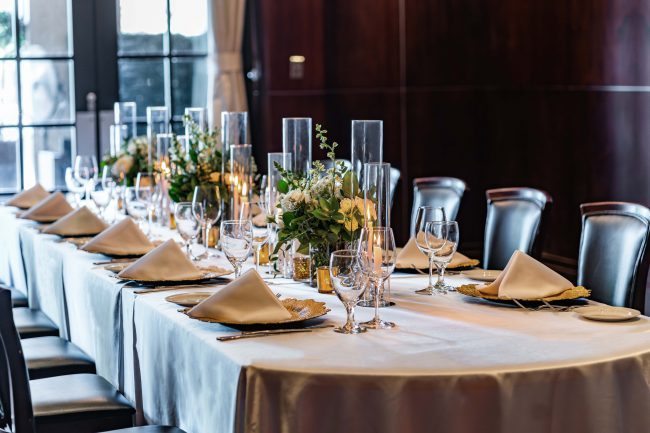 Truluck's Woodlands Naples room view of the boardroom table with the sun shining through the large glass window doors onto a table dressed in glass candleholders, fresh florals, gold charger plates and satin napkins.