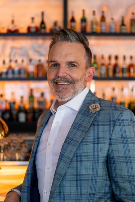 headshot of Riley Hutton at the Austin downtown location standing in front of the bar with a white shirt and blue plaid jacket with a flower pin