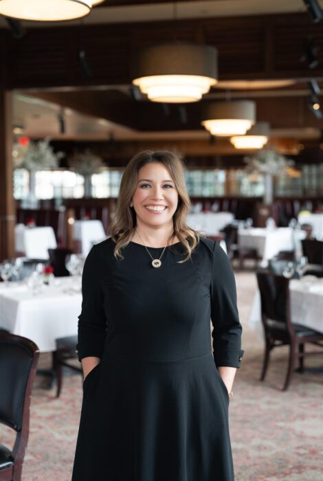 Amber Scheer, director of sales and events, smiles confidently inside Truluck’s elegant dining room, dressed in black, with white-tablecloth tables and soft ambient lighting behind her.