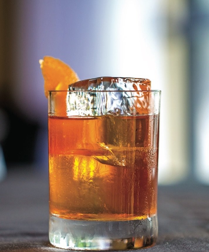 Old fashioned poured in a cocktail glass with a large block of ice and an orange rind