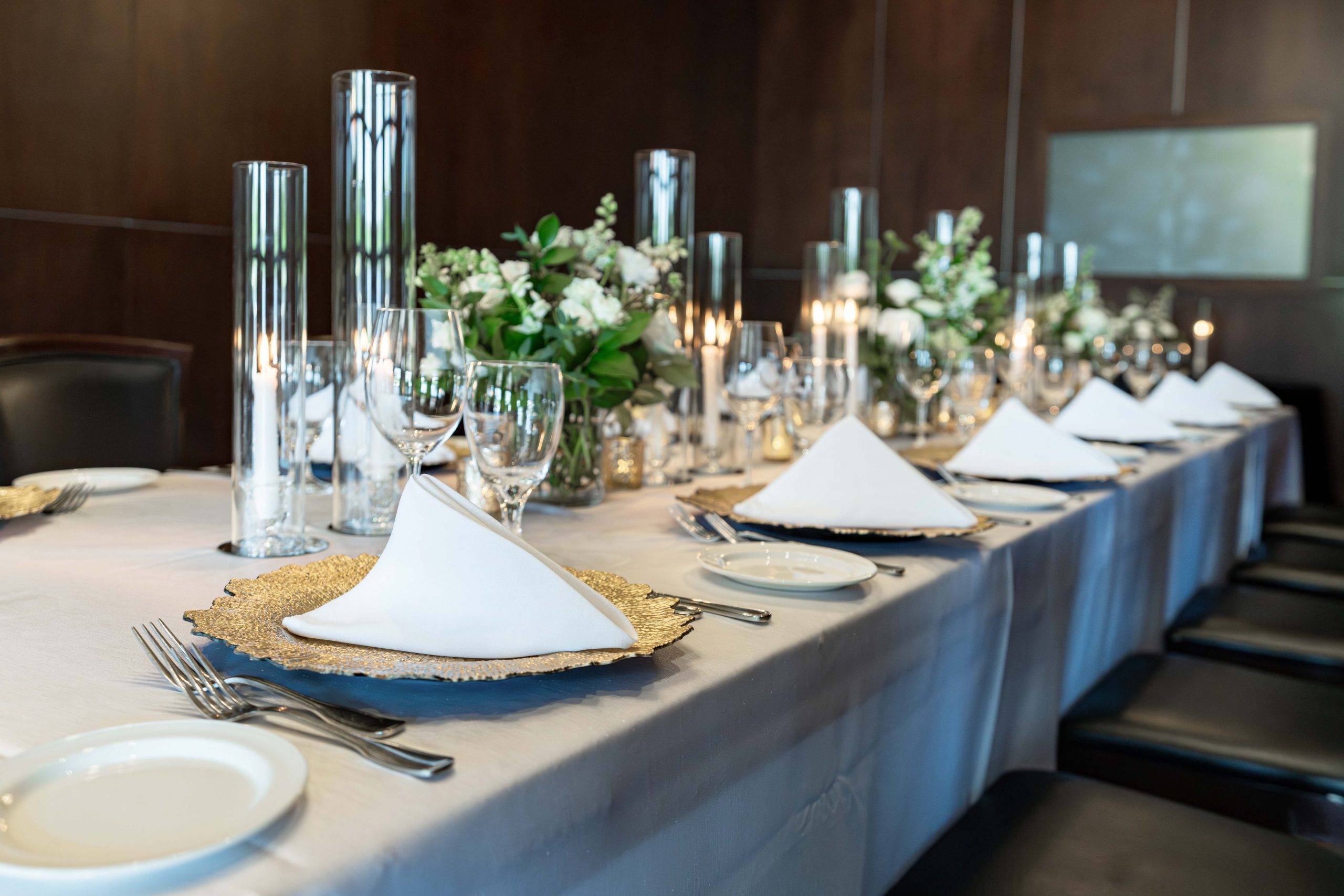 A boardroom style table view from the corner end, the table is dressed with gold chargers, silverware, water and wine glasses, gold tapered candles and white flowers