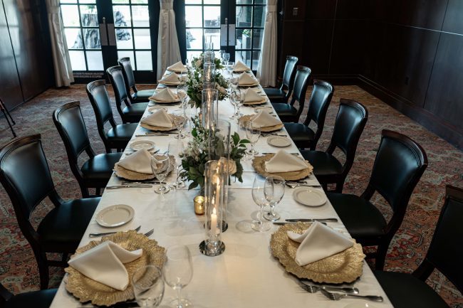 The Naples room at the Woodlands location set up with a long rectangular table dressed in white linen, flowers, glass candles and gold chargers with glassed in French doors overlooking the terrace.