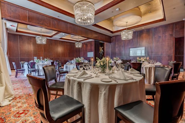The Stone Crab Room at The Woodlands location. Round tables draped in white linen table clothes, dressed with full place settings and surrounded by mahogany wood chairs. Ceilings adorned with crystal chandeliers.