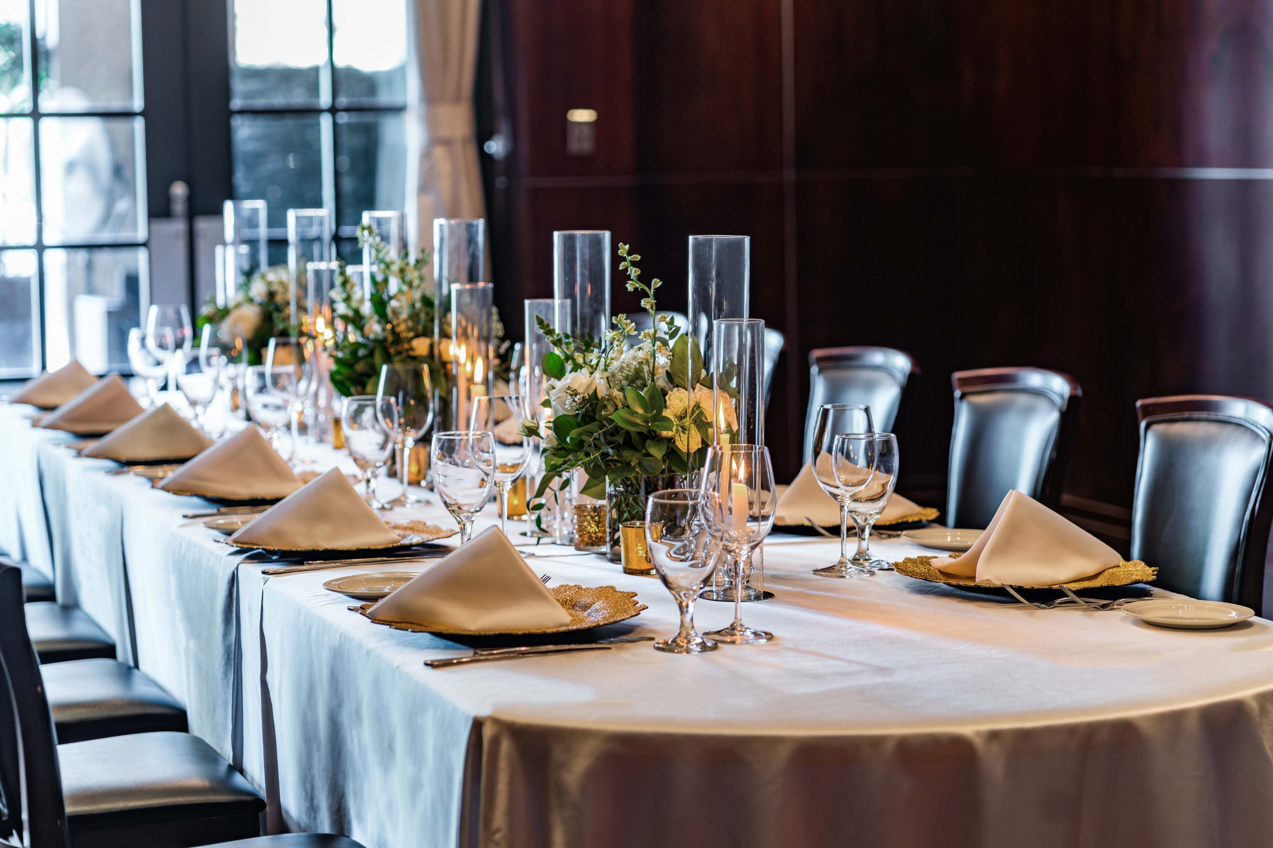 a rectangular table setup with gold chargers, white napkins, fresh flowers, cylinder glass candle holders