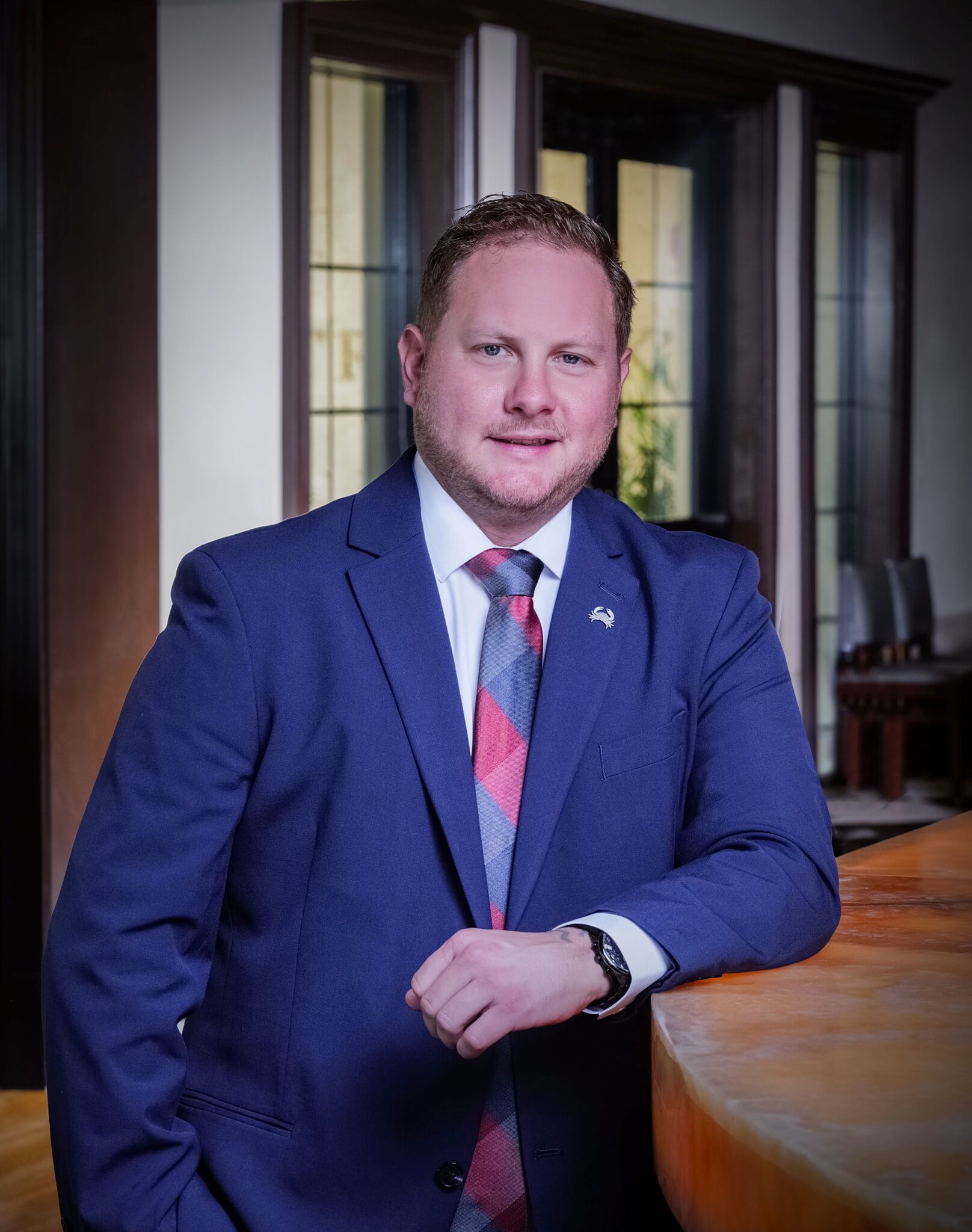 headshot of Johnny Griesedieck in the Naples bar area with his elbow propped on the bartop. He's in a blue suit, white button down shirt and multi colored tie