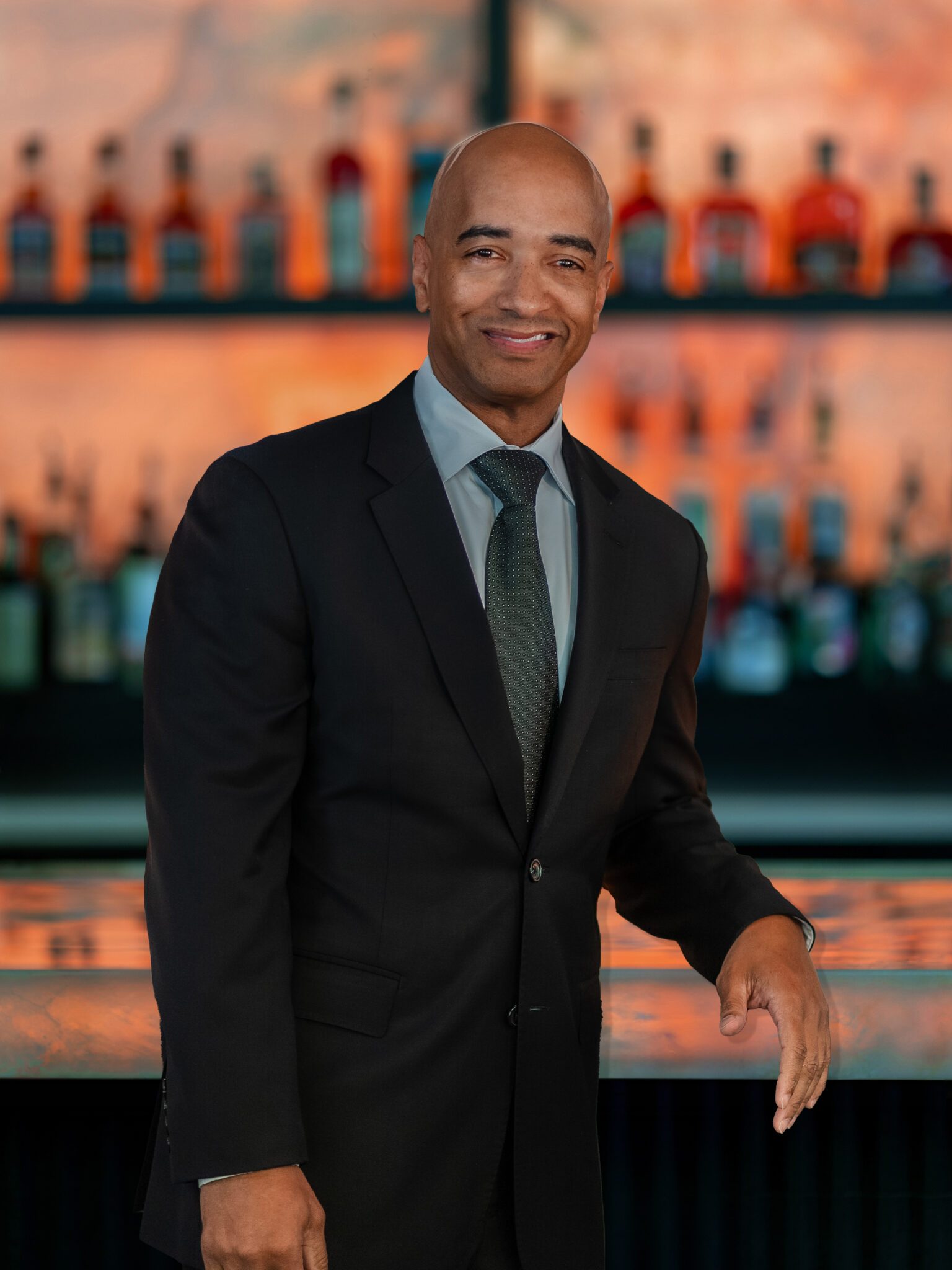 headshot of DC general manager, Anthony Ray, in a dark suit with shirt and tie standing in front of the glowing bar top with his elbow on the bar