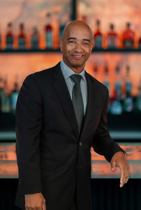 headshot of DC general manager, Anthony Ray, in a dark suit with shirt and tie standing in front of the glowing bar top with his elbow on the bar