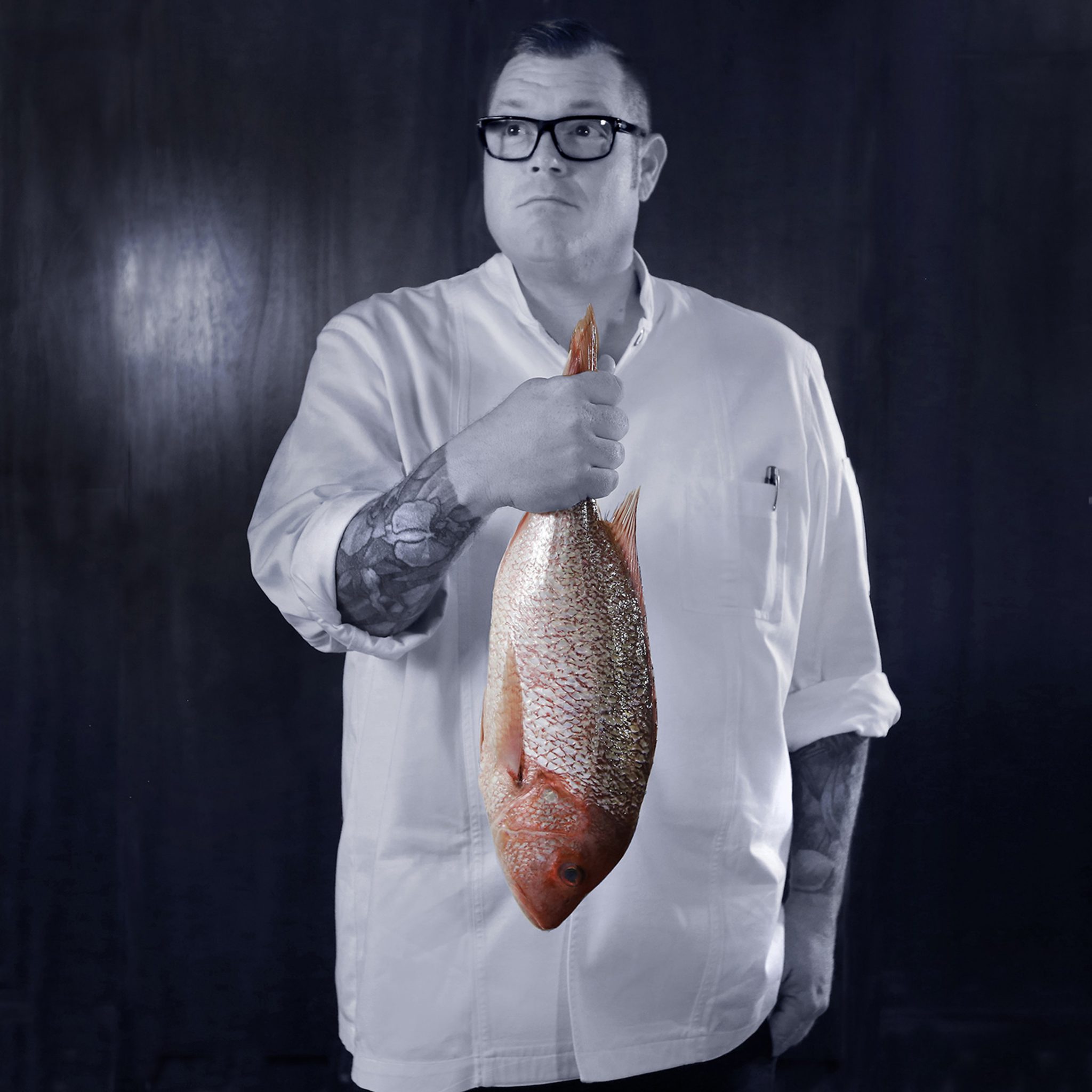 picture of Director of Culinary, Chef Brian Wubbena in a black and white photo holding a fresh fish that is in the color orange.