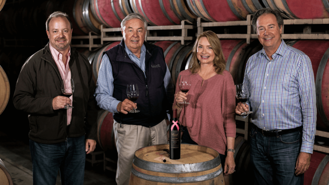 picture of the J. Lohr family holding wine glasses with wine casks in the background