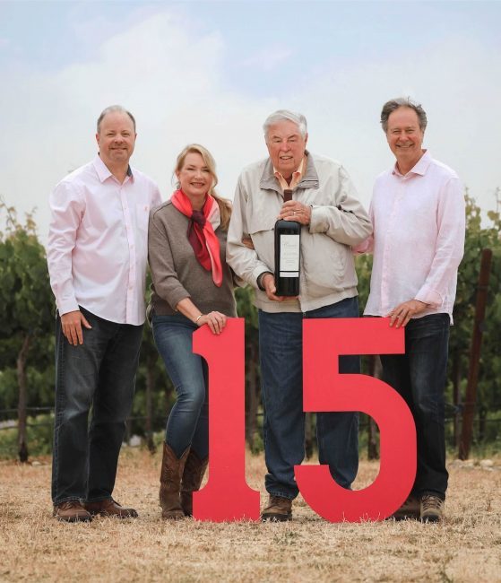 Lohr family photo with large 3' tall number 15 in front and them holding Carol's Vineyard bottles in front of the vineyards 