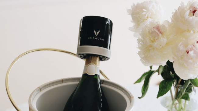 Picture of a bottle of champagne with the Coravin wine sparkling cap on top of the bottle. The bottle is in a chilling bucket with a vase full of white peonies on the right.