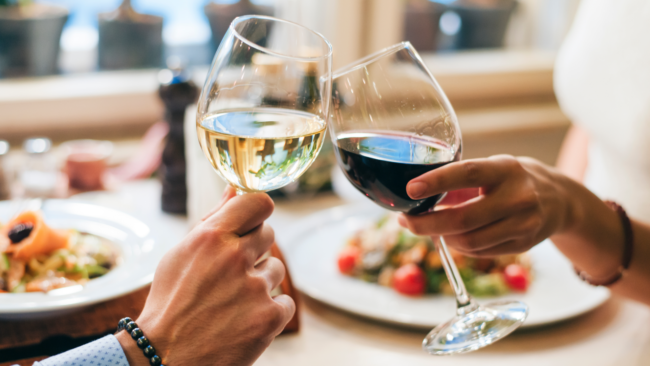 picture of a glass of red and white wine clinking together over a table of food