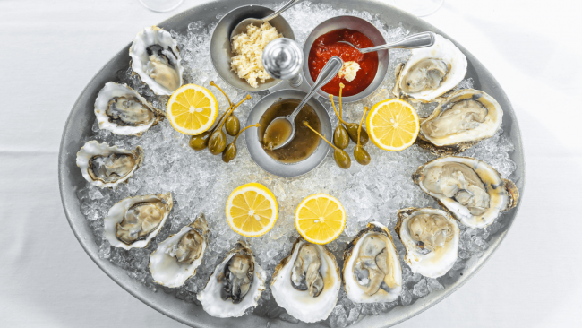 a platter of freshly shucked oysters on a bed of crushed ice with a lemon half, caper berries, horseradish, cocktail sauce and champagne mignonette