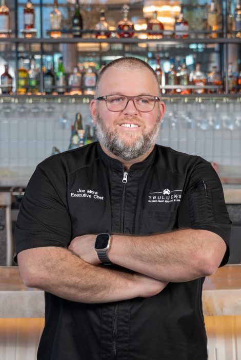 headshot of Chef Joe Mora in a black logo chef coat with his arms folded across his chest in from of the bar