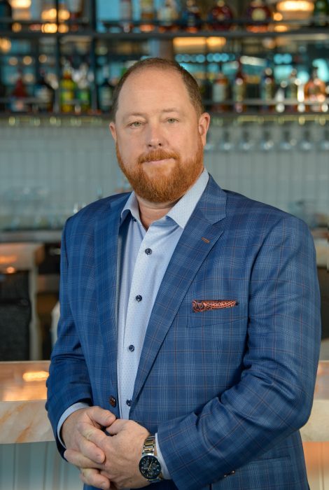 headshot of operating partner Todd Perry in a blue plaid suit with his hands crossed and leaning on the bar top