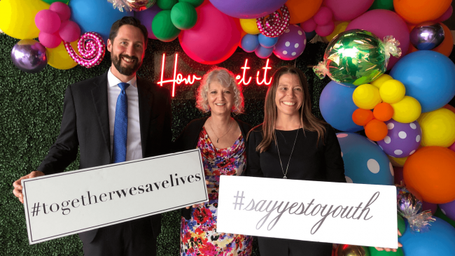 photo of Truluck's staff with #togetherwesavelives and #yestoyouth signs in front of a balloon backdrop