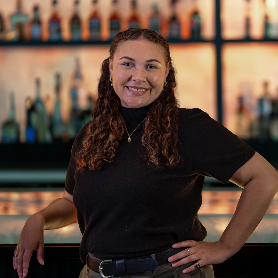 headshot of Kyla Adams, Truluck's Houston sales & events manager. She is standing poised with one elbow on the bar top and her opposite hand on her hip
