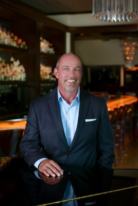 headshot photo of general manager, Ken Allen, in the bar at the Naples location with his hand on the piano