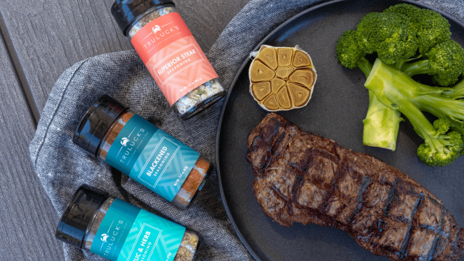 photo of garlic & herb seasoning, superior steak seasoning and blackened seasoning in a labeled spice bottle next to a plate with a New York strip that is grilled, steamed broccoli and a garlic halve