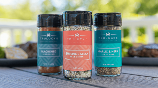 photo of blackened seasoning, superior steak seasoning and garlic & herb seasoning in a labeled spice bottle on a table out side with steaks in the background