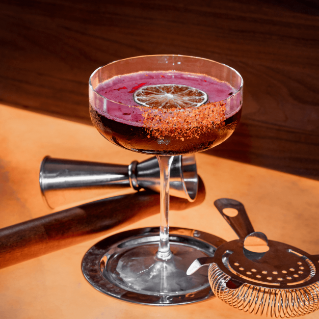 picture of the Smoke in the Garden cocktail with a dried lime wheel and tahin rim with a strainer, jigger and muddler in the background