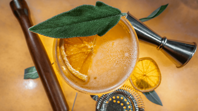 photo of the Backward Glance cocktail topped with a mint leaf with a jigger, muddler and strainer and lemon in the background
