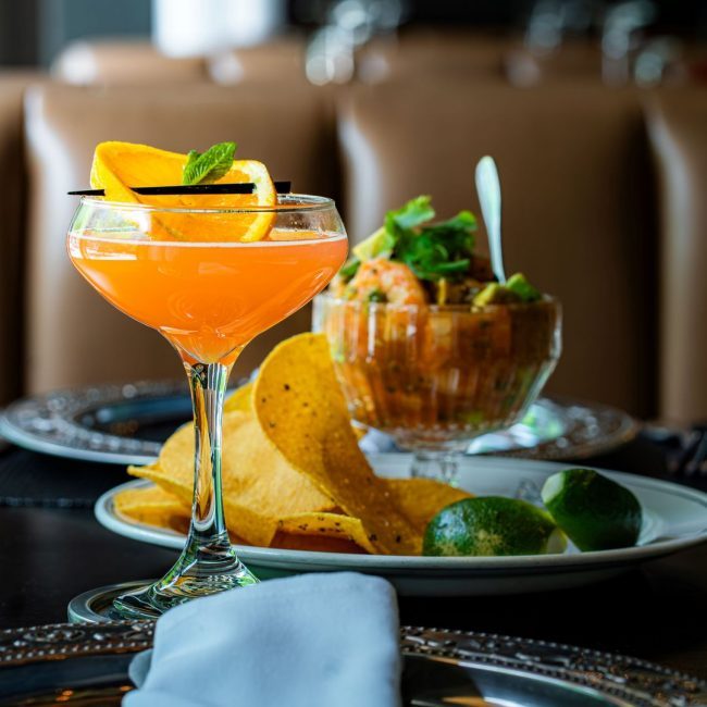 High flier cocktail with an orange wheel garnish and a shrimp campechana appetizer in a glass bowl with chips and lime in the background