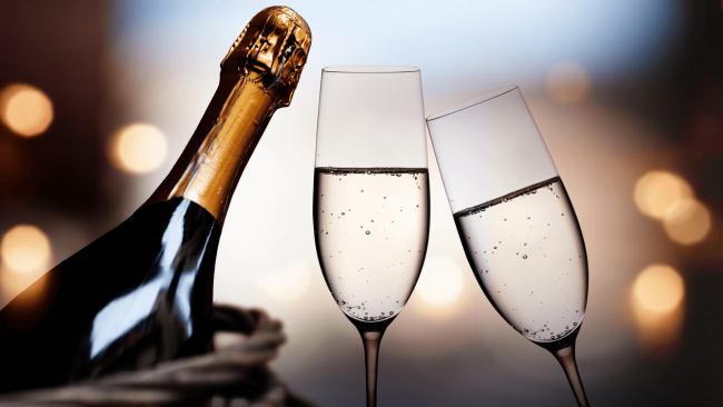 photo of a bottle of champagne that is unopened next to 2 glasses of champagne. One glass is tipped toward the other glass and the background has the bokeh effect.