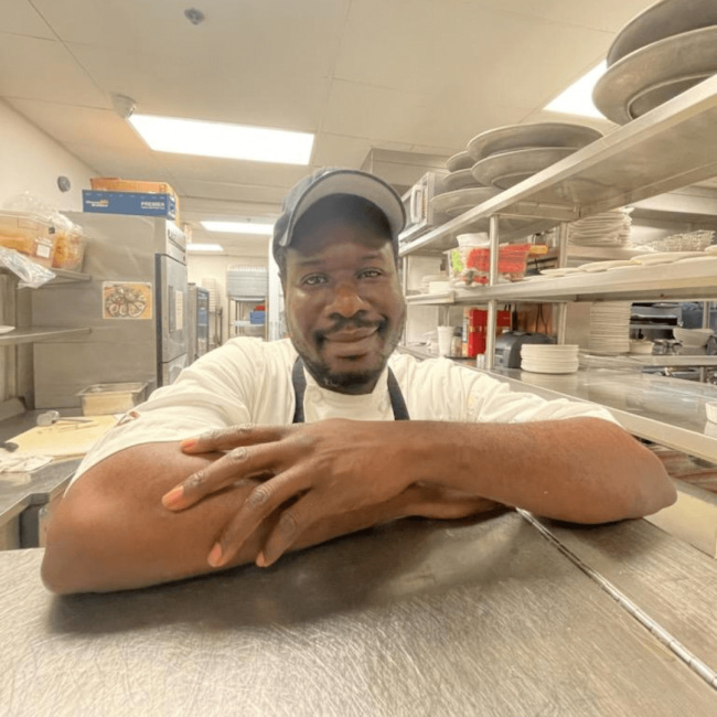 photo of line cook, Andrew on the main kitchen line in a hat and apron with his elbows up on the table