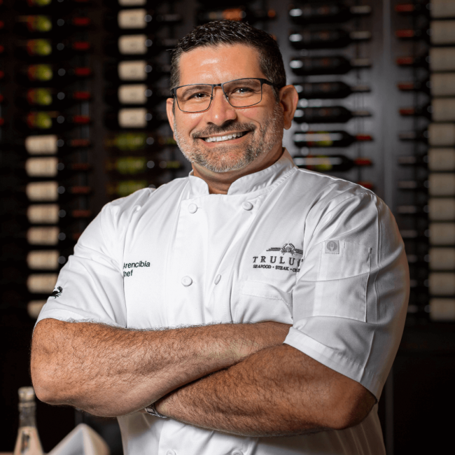 chef Odel Arencibia in his chef coat in front of the wine wall in Miami